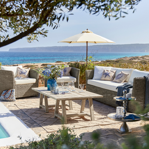 Gartenmöbel-Set aus grauem Technorattan mit cremefarbenen Kissen und Sonnenschirm vor Meerblick