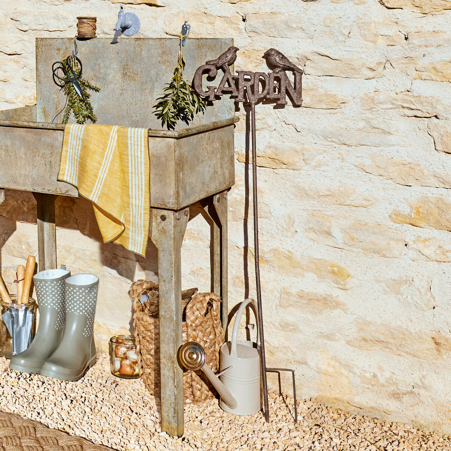 Gartenstab Girra Metall-Gartenspüle mit gelbem Tuch und Gartengeräten vor Steinmauer im Außenbereich