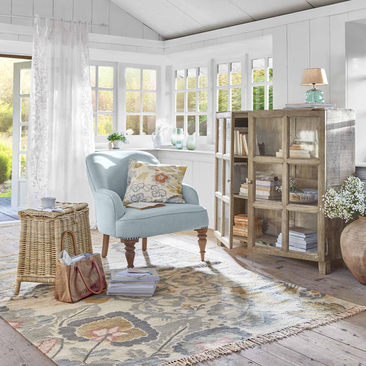 Gemütliches Zimmer mit hellblauem Sessel, Holzvitrine und floralem Teppich im Landhausstil