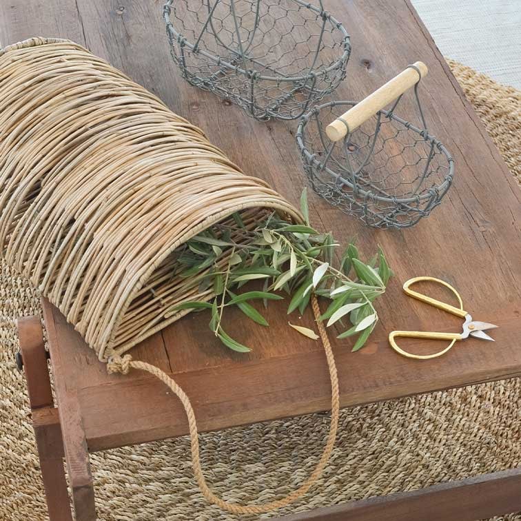 Weidenkorb mit Kordelgriff auf Holztisch mit grünen Zweigen und goldfarbenen Gartenscheren
