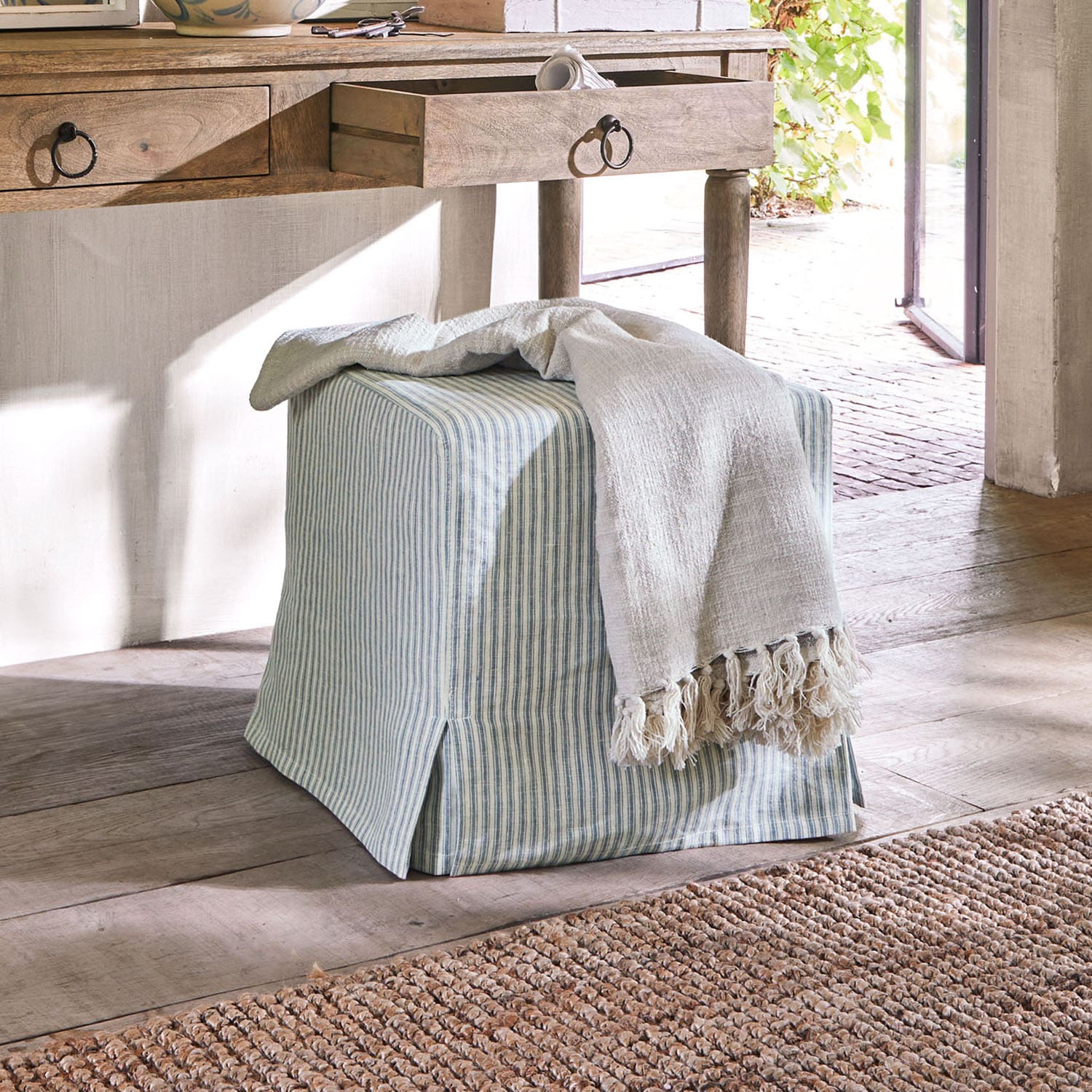 Quadratischer Pouf aus Baumwolle mit blau-weiß gestreiftem Bezug und hellem Fransenplaid auf Holzboden