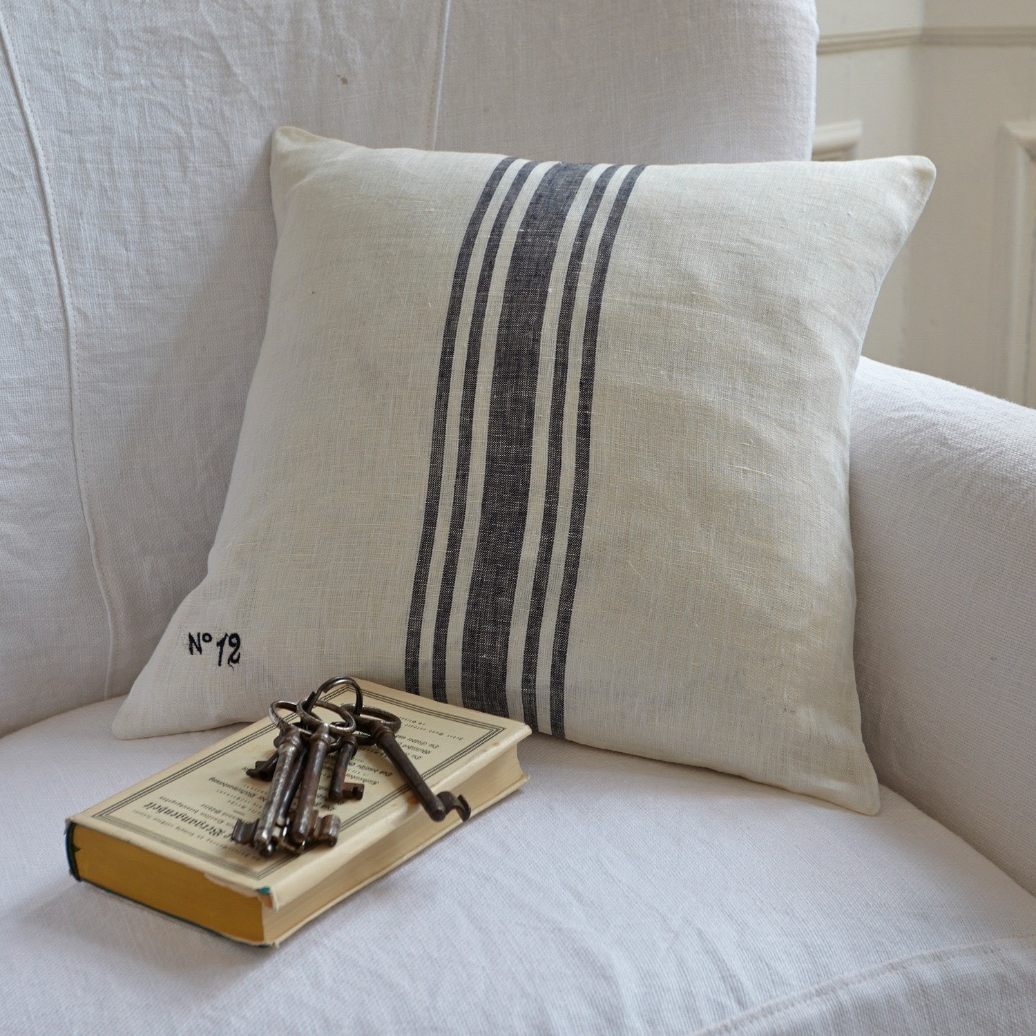 Kissen Lucian Dekoratives Kissen aus Leinen in Creme mit schwarzen Streifen auf weißem Sofa mit Buch und Schlüsseln