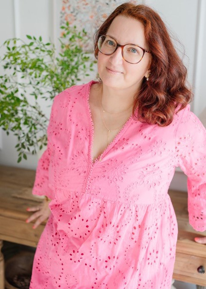 Frau in rosa Lochstickerei-Kleid mit zartem Muster, lehnt an Holztisch mit Pflanze im Hintergrund
