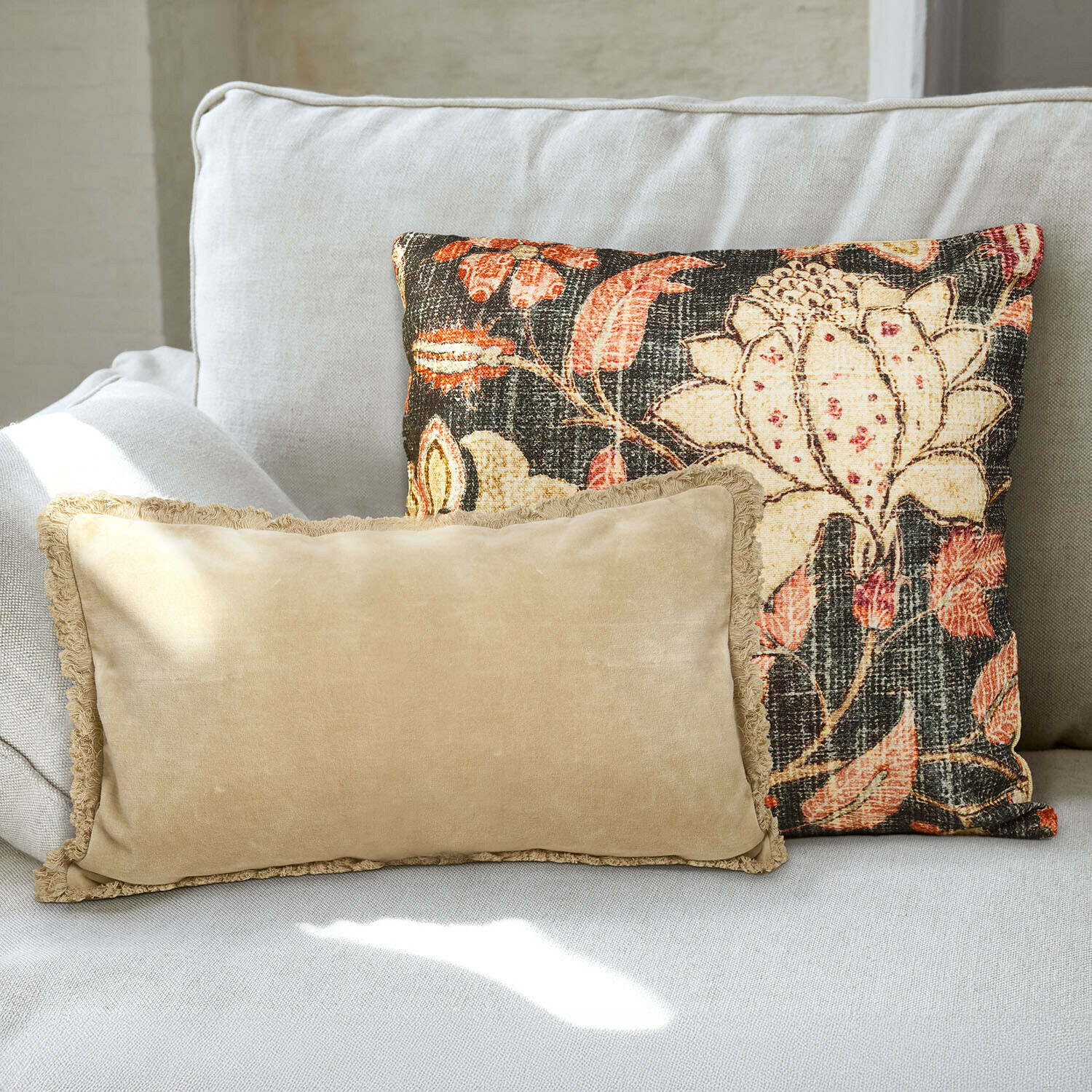 Kissenhülle 2er Set Ambrayelle Zwei dekorative Kissen auf hellgrauem Sofa, eines beige mit Fransen, das andere mit buntem Blumenmuster
