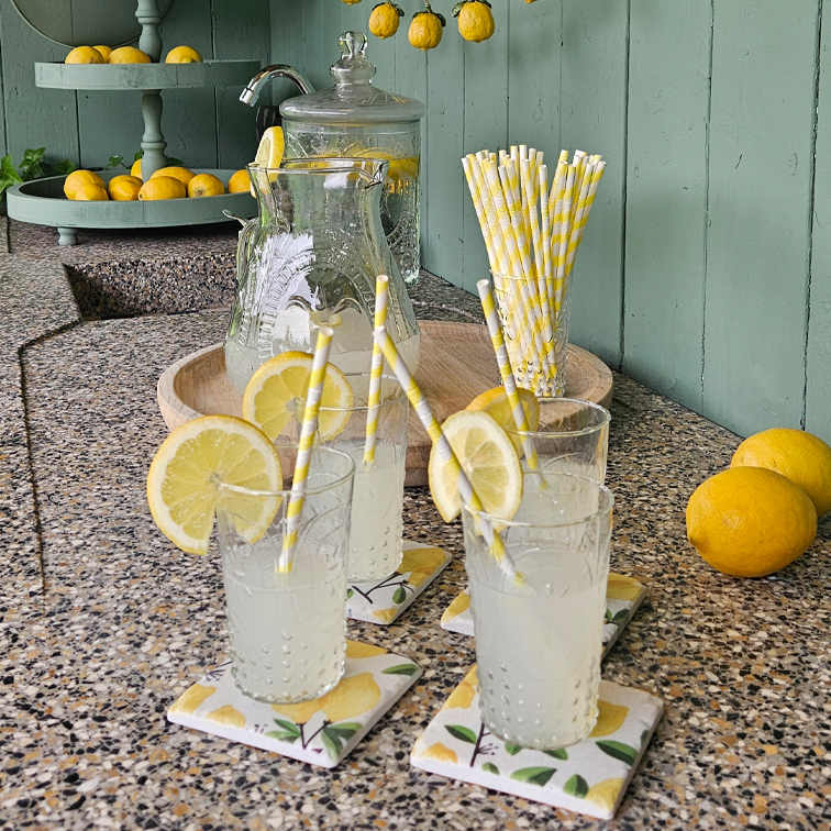 Gläser mit Limonade und Zitronenscheiben auf Steinplatte mit Papierstrohhalmen und Tablett mit Zitronen