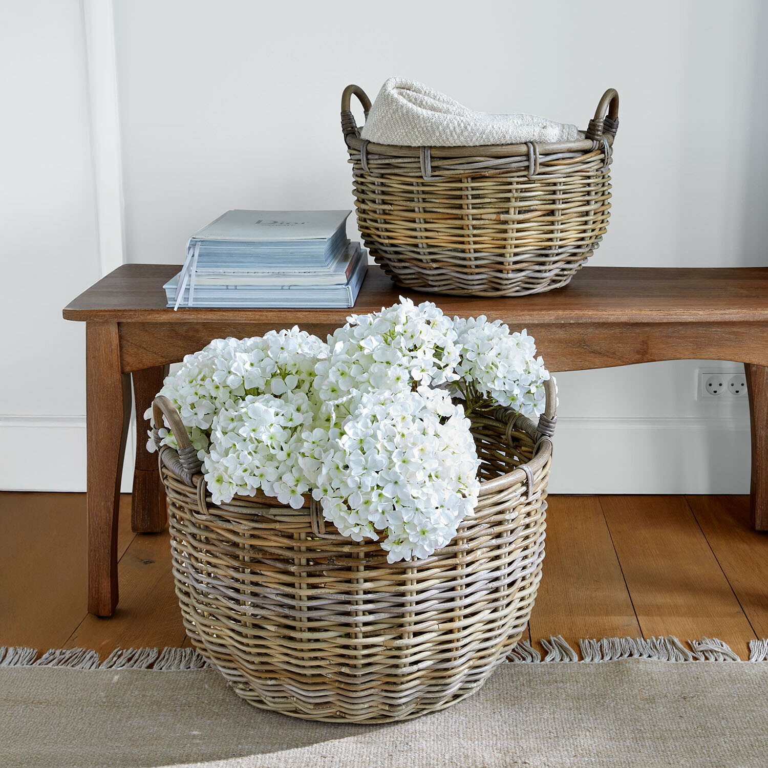 Großer Korb aus Weide mit weißen Blumen und kleinerer Korb mit Decke auf Holzbänkchen im Wohnzimmer