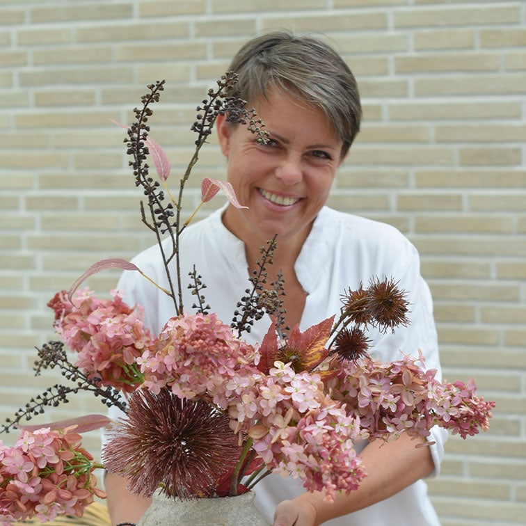 Frau in weißer Bluse hält Vase mit rosa und braunen getrockneten Dekoblumen