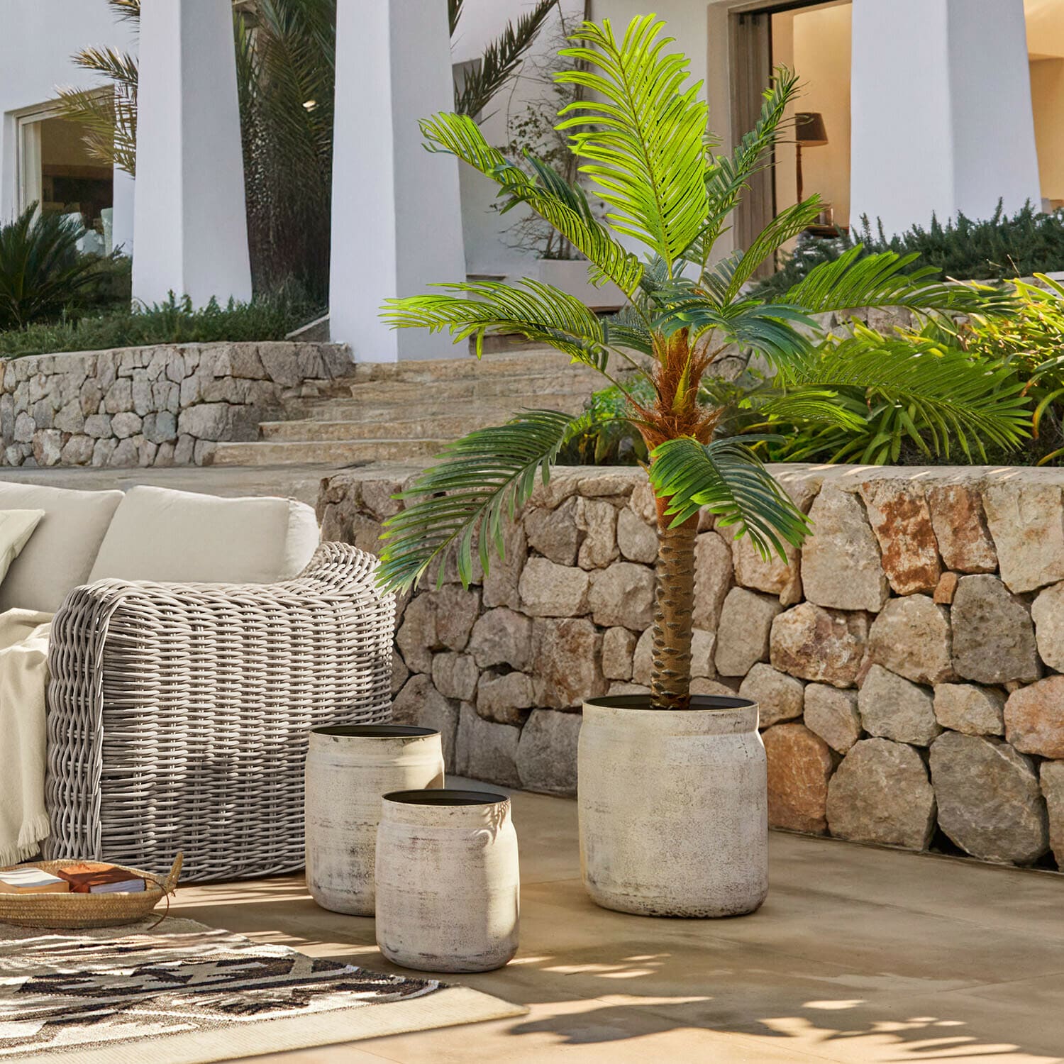 Set aus drei hellen Betontöpfen auf Terrasse mit grüner künstlicher Palme und Rattanmöbeln