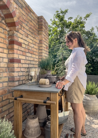 Frau in weißem Hemd und beigen Shorts steht an einem Holztisch mit Betonplatte im Garten vor einer Ziegelwand und Pflanzen