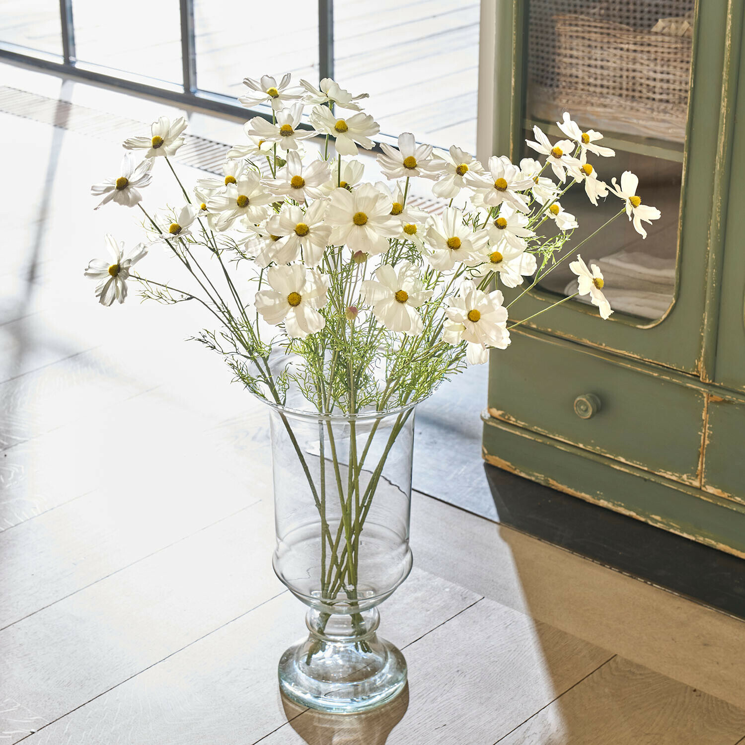 Transparente Glasvase mit weißen Kosmosblumen auf hellem Boden im natürlichen Licht