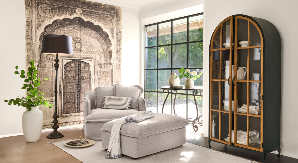 Gemütliches Wohnzimmer mit beigem Sessel und Hocker, Metallvitrine mit Holztüren und dekorativem Wandteppich