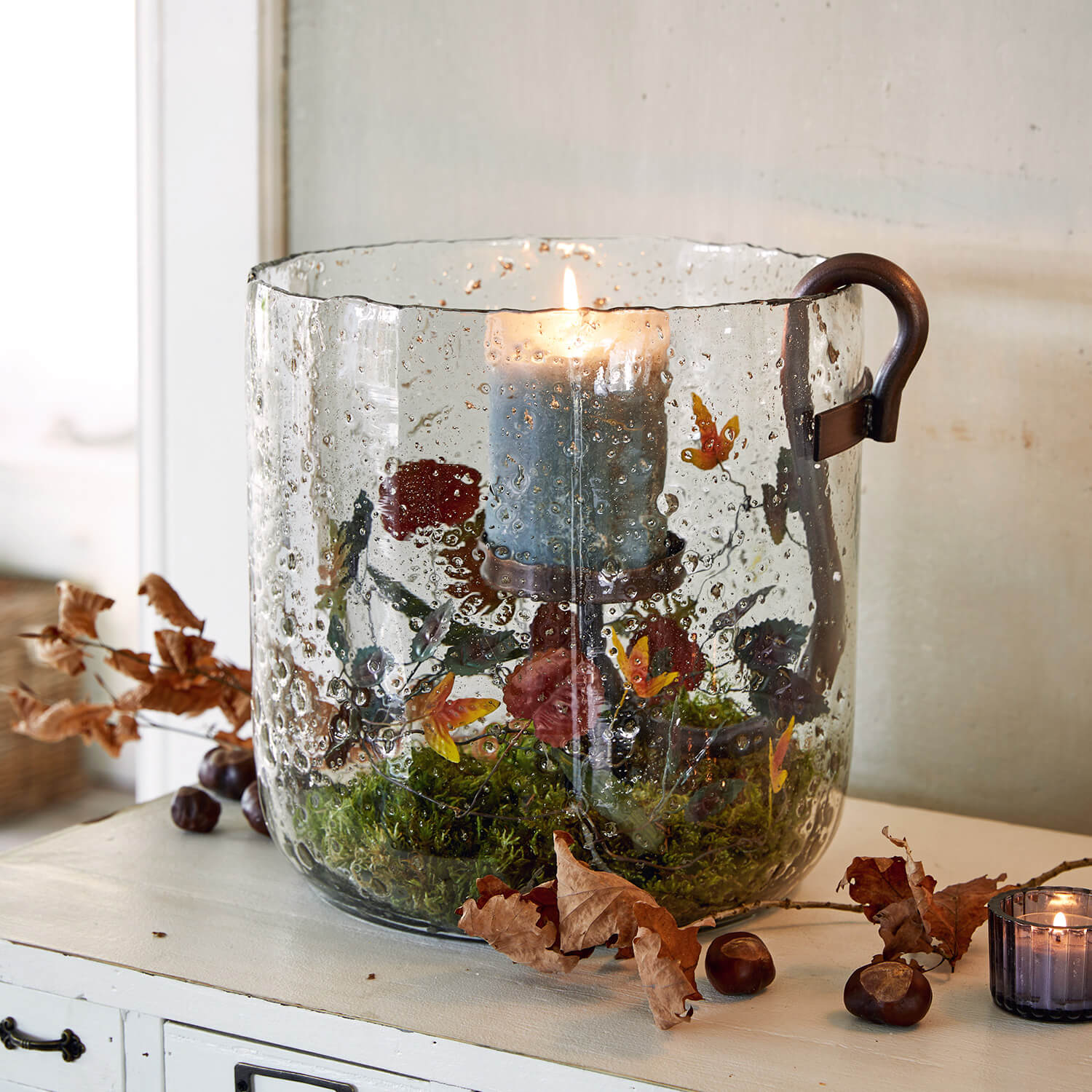 Glaswindlicht mit blauer Kerze, floralem Muster und herbstlicher Dekoration aus Blättern und Moos