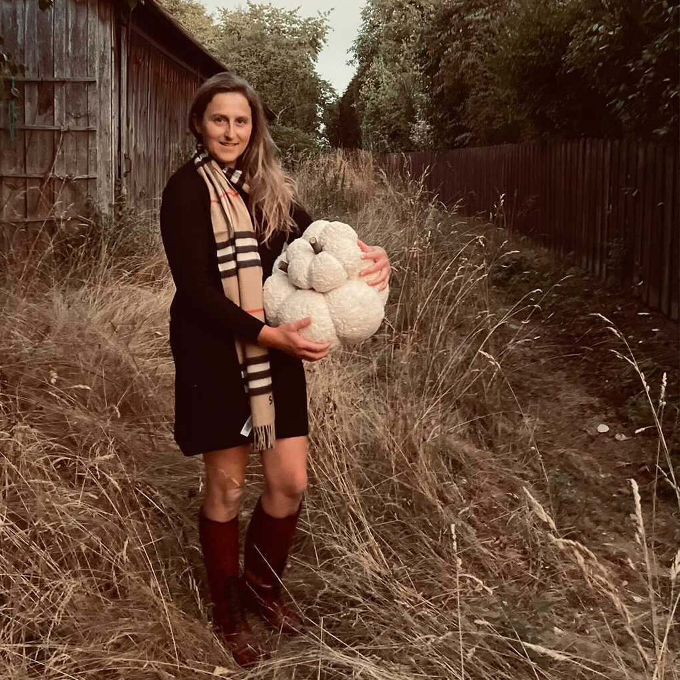 Frau in schwarzem Kleid und Schal hält weißen Dekokürbis auf herbstlicher Wiese