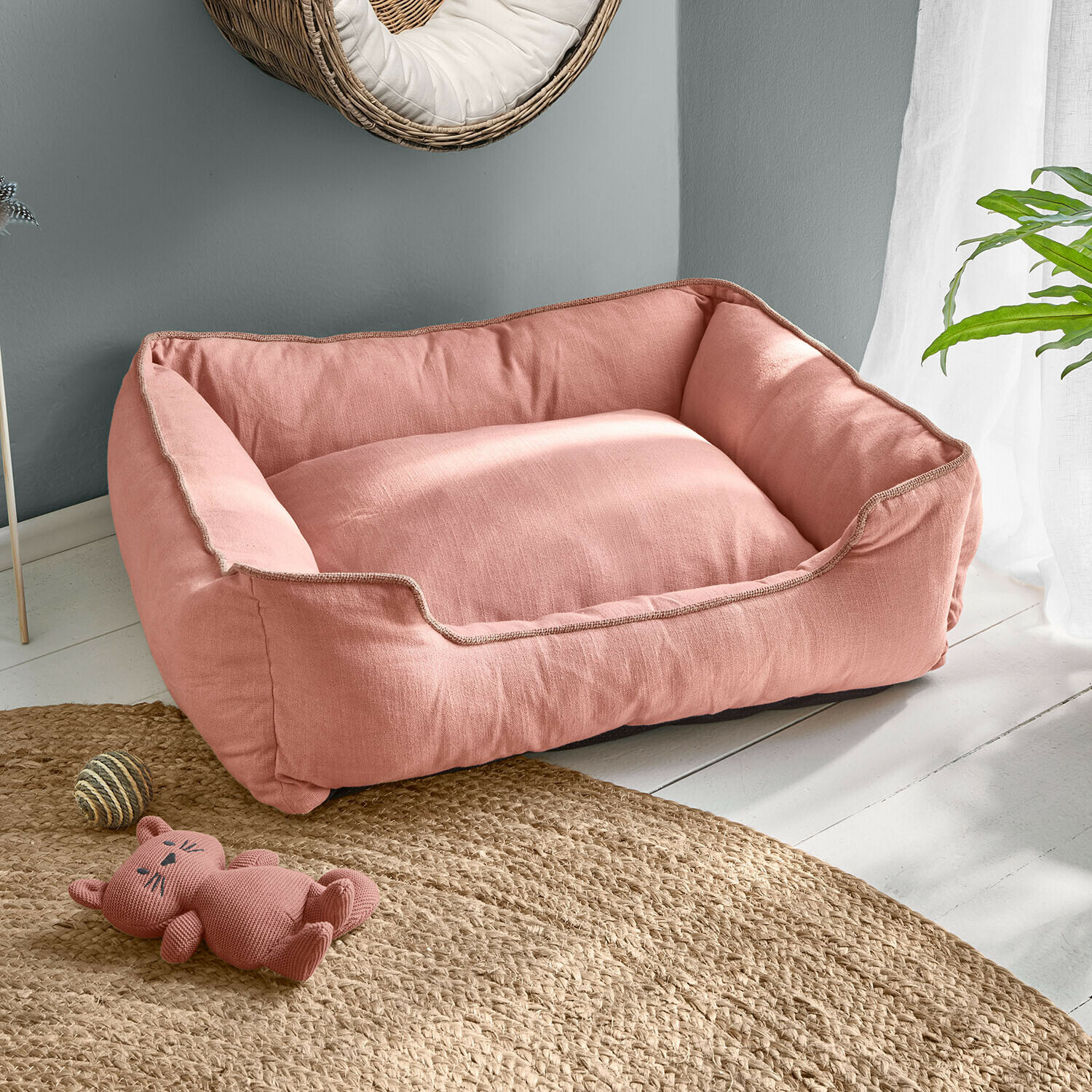 Rosa Hundebett aus weichem Stoff auf hellem Boden mit Katzen-Spielzeug und Jute-Teppich