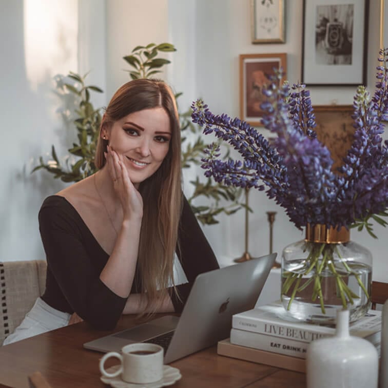 Frau arbeitet am Laptop an einem Holztisch mit lila Blumenstrauß und Kaffeetasse