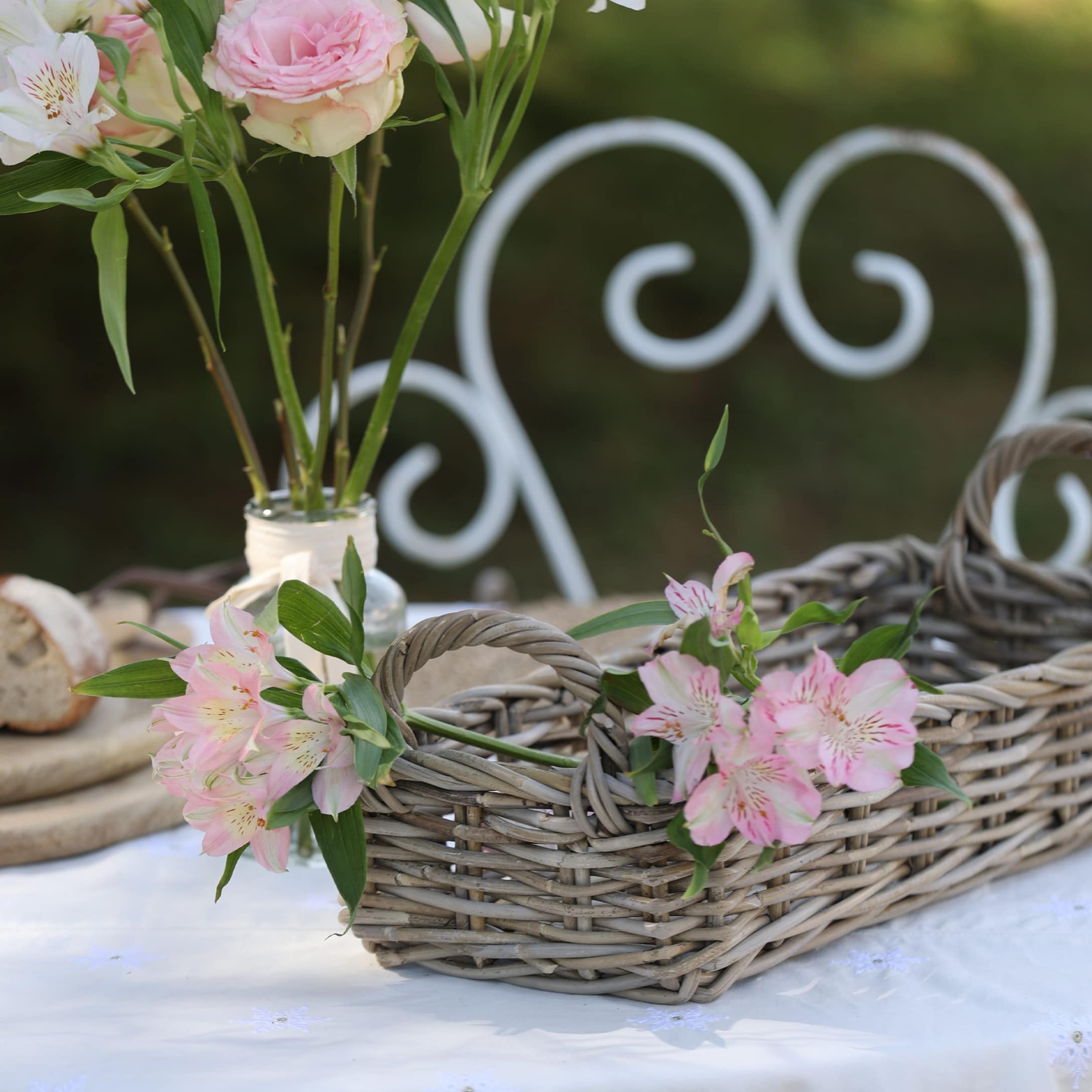 Rustikaler grauer Weidenkorb mit rosa Blumen auf weißer Tischdecke im Garten