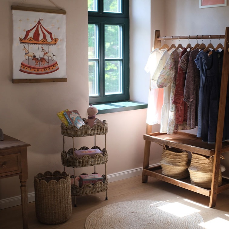 Dreistöckiges Korbregal mit Spielzeug und Kinderkleidung an einem Holzkleiderständer in hellem Raum