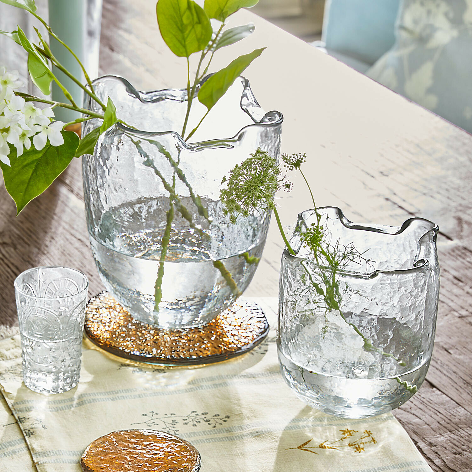 Transparente Glasvasen mit welligem Rand auf Holztisch mit grünen Pflanzen und Dekoration