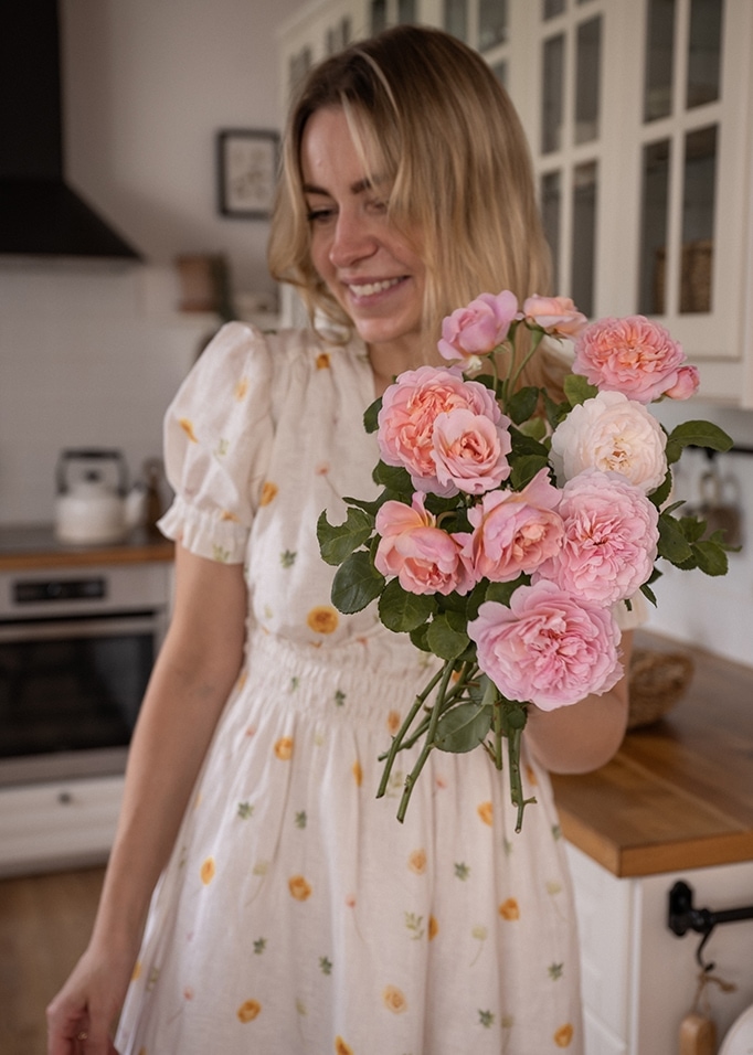 Frau im weißen Blumenkleid hält einen Strauß rosa Rosen in moderner Küche