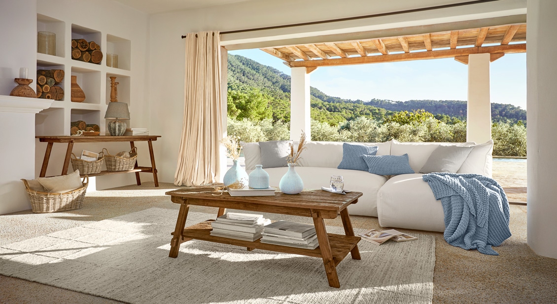 Gemütliches Wohnzimmer mit weißem Sofa, Holztisch und blauen Kissen mit Blick auf grüne Hügel