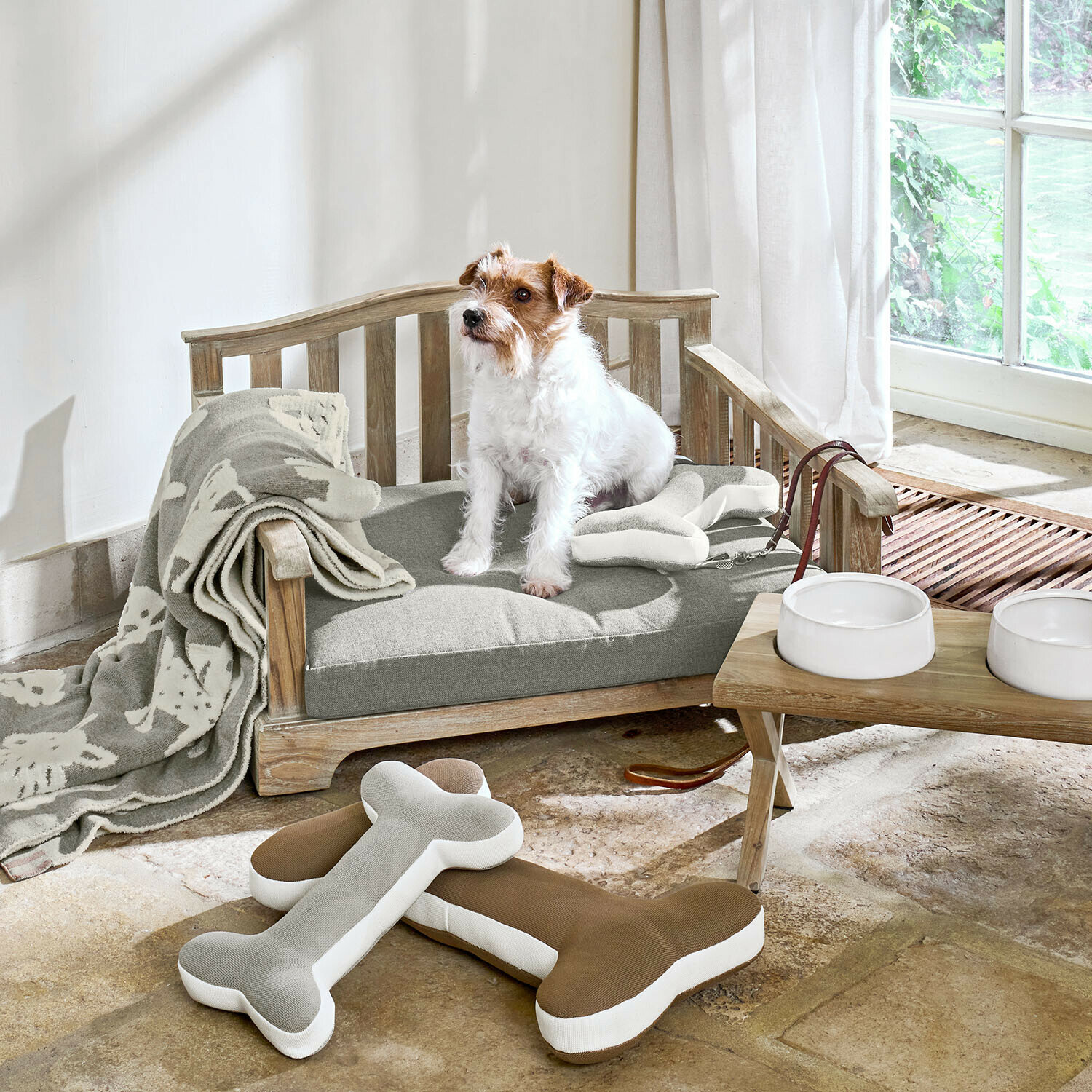 In- & Outdoor-Haustiersofa Waggins Holz-Hundebett mit grauem Kissen, Knochen-Spielzeug, helle Decke und weißen Futternäpfen am Fenster
