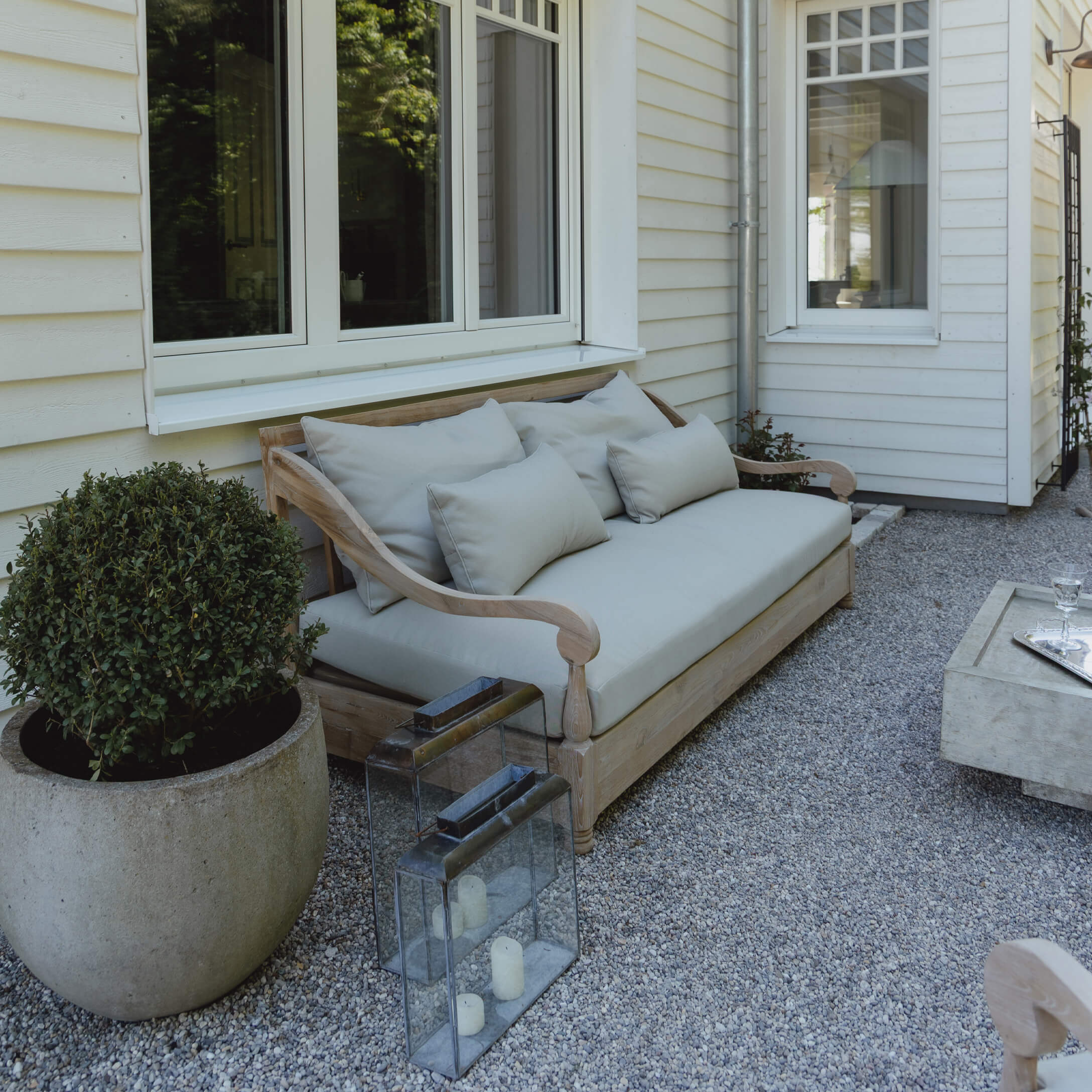Holz-Gartensofa mit hellgrauen Kissen auf Kiespatio vor weißem Haus