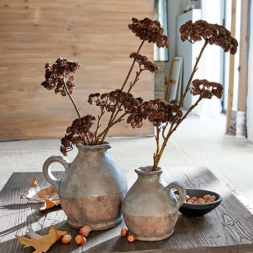 Zwei graubraune Keramikkrüge mit getrockneten Blumen auf einem Holztisch mit Haselnüssen