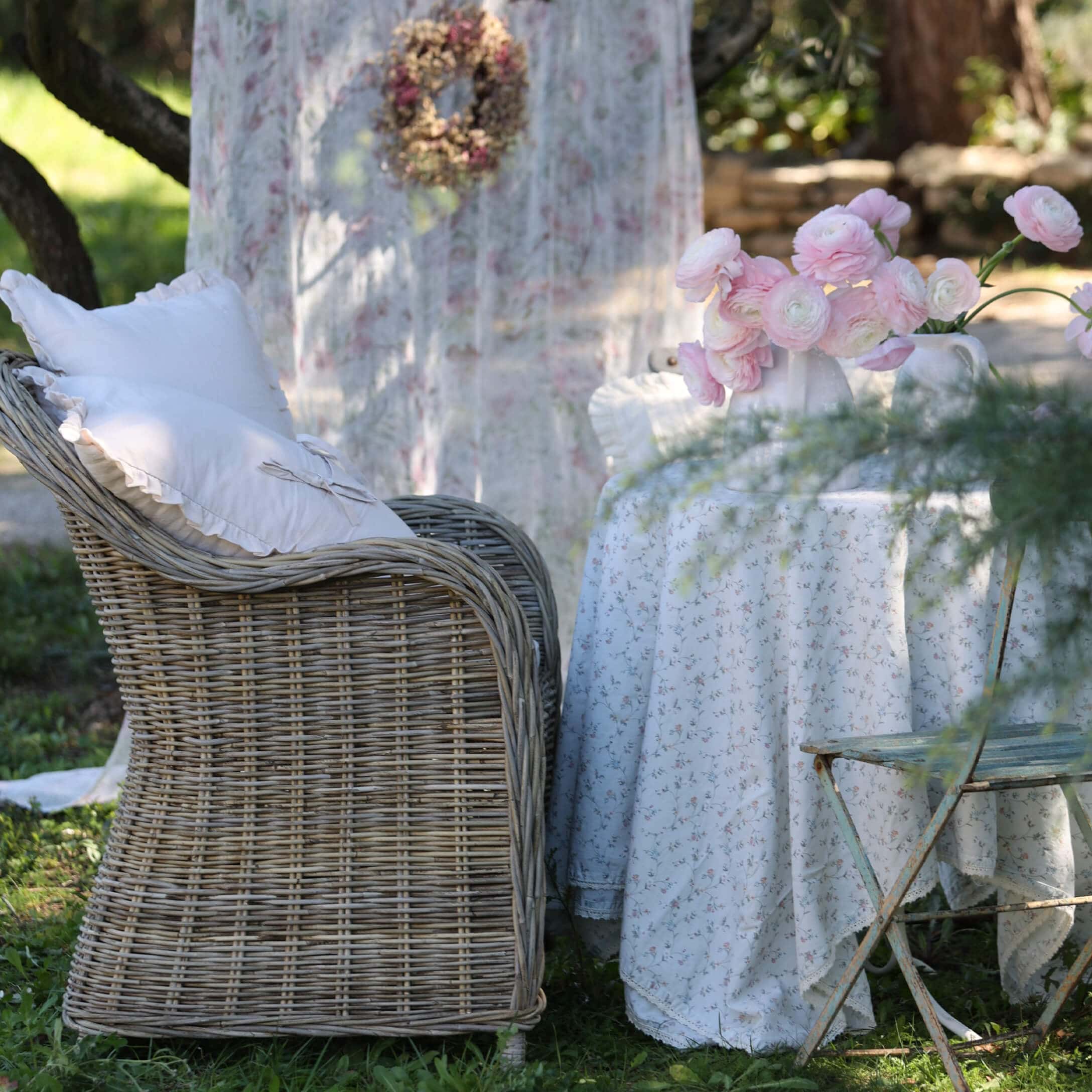 Wiklinersessel mit weißen Kissen neben einem Tisch mit floralem Tischtuch und rosa Blumen im Garten