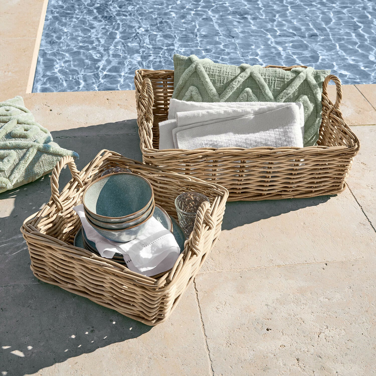 Zwei geflochtene Körbe mit Griffen, gefüllt mit grünen Kissen und weißen Handtüchern am Poolrand