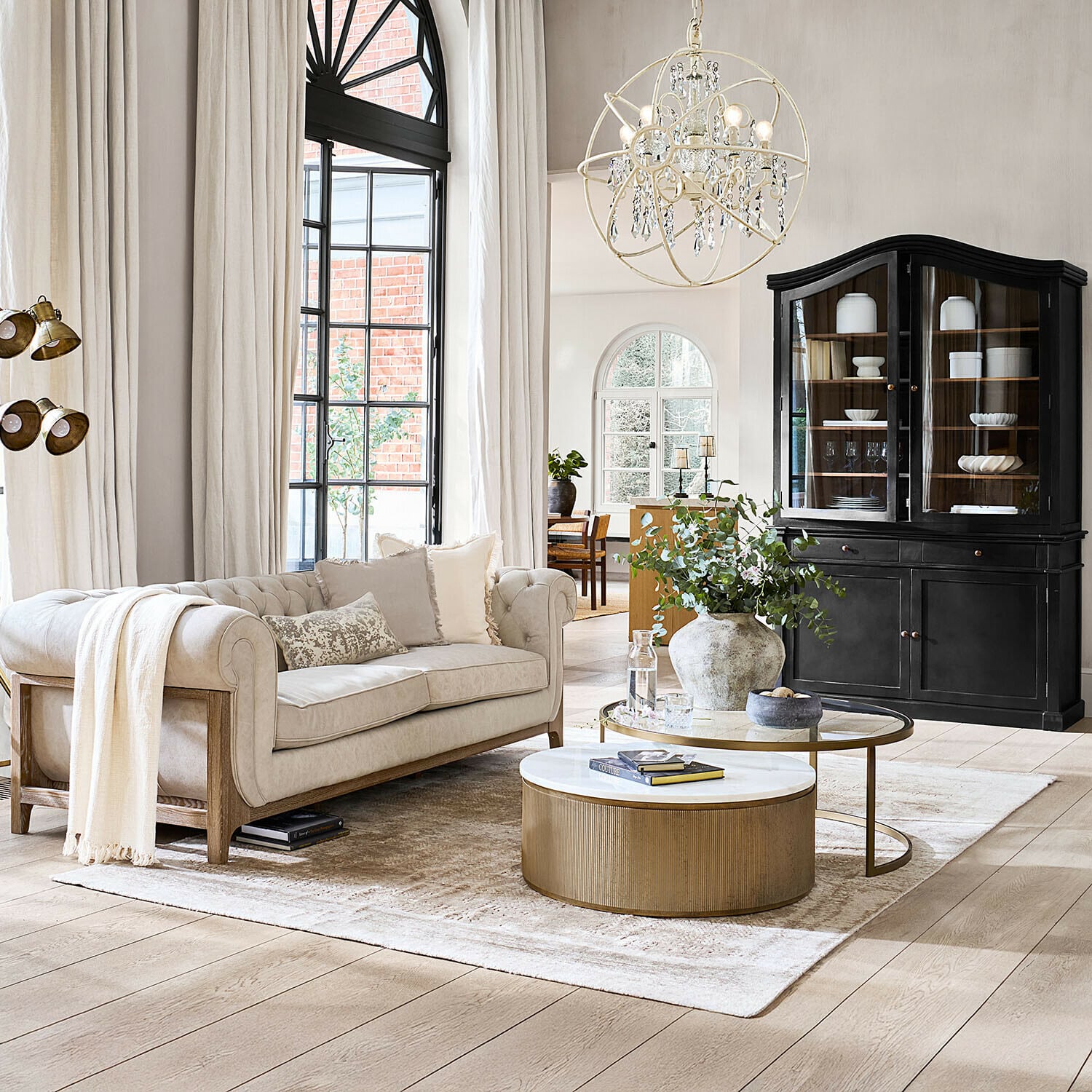 Teppich Ashmere Gemütliches Wohnzimmer mit beigem Sofa, goldfarbenem Couchtisch und schwarzem Vitrinenschrank vor hellen Vorhängen