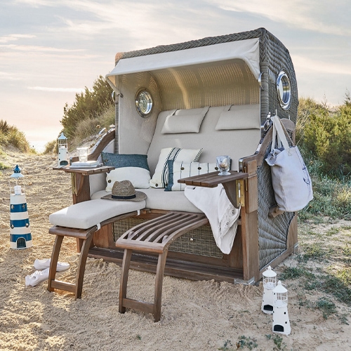 Strandkorb aus Rattan mit beigen Polstern und Holzfußstützen im Sand, ideal zum Entspannen