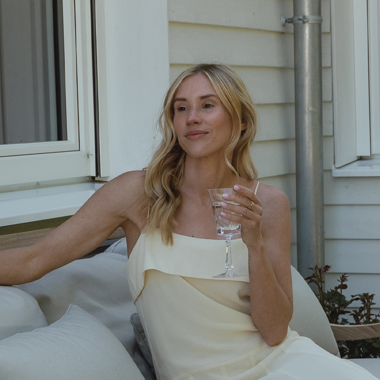 Frau in cremefarbenem Kleid entspannt draußen mit Glas in der Hand auf Garten-Couch