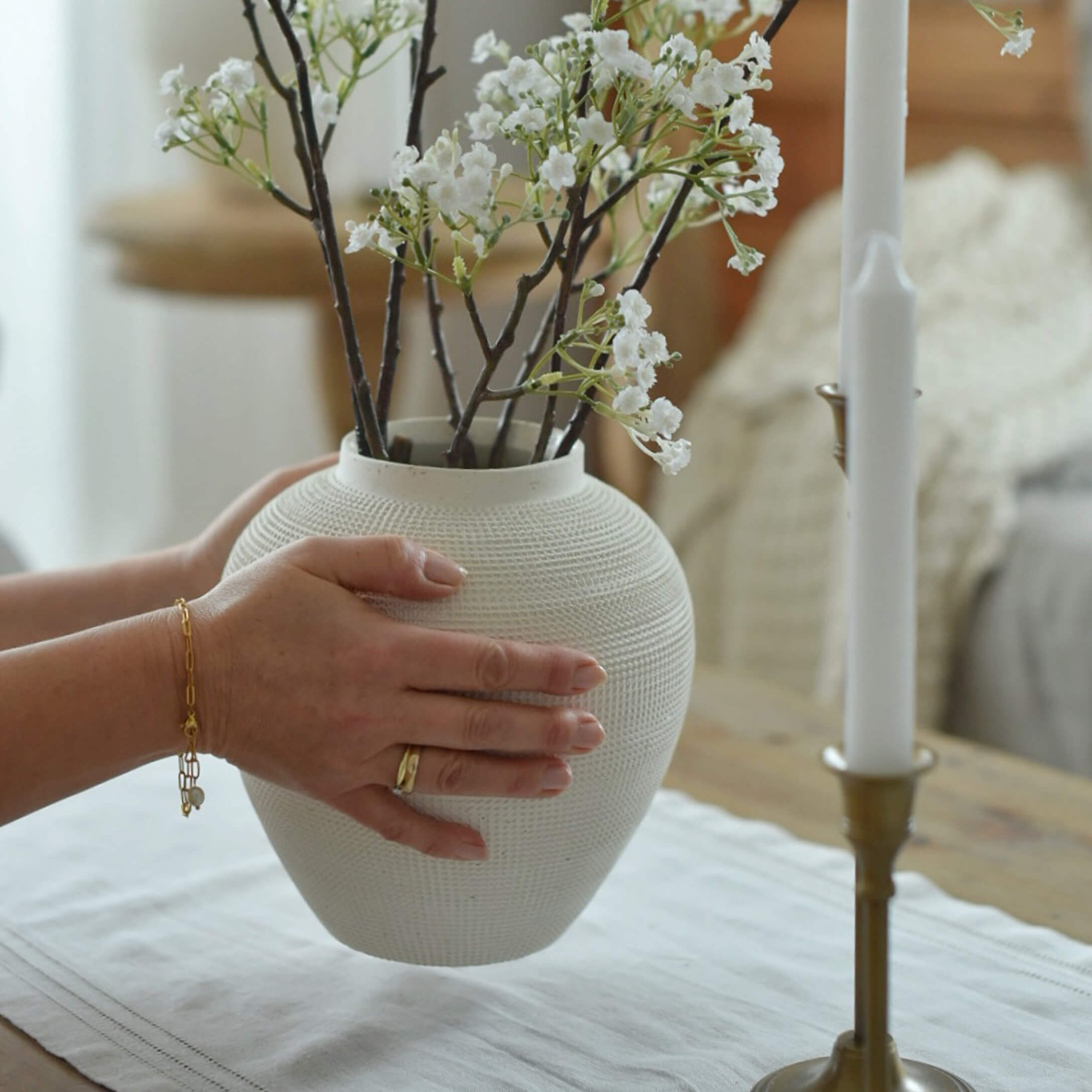 Weiße strukturierte Keramikvase mit weißen Blütenzweigen, gehalten von Händen auf einem Tisch