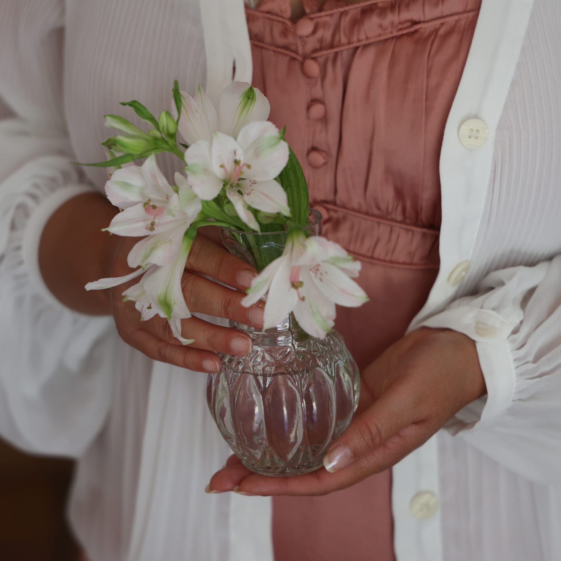 Glasvase mit weißen Blumen, gehalten von Person in weißem Cardigan und rosa Kleid