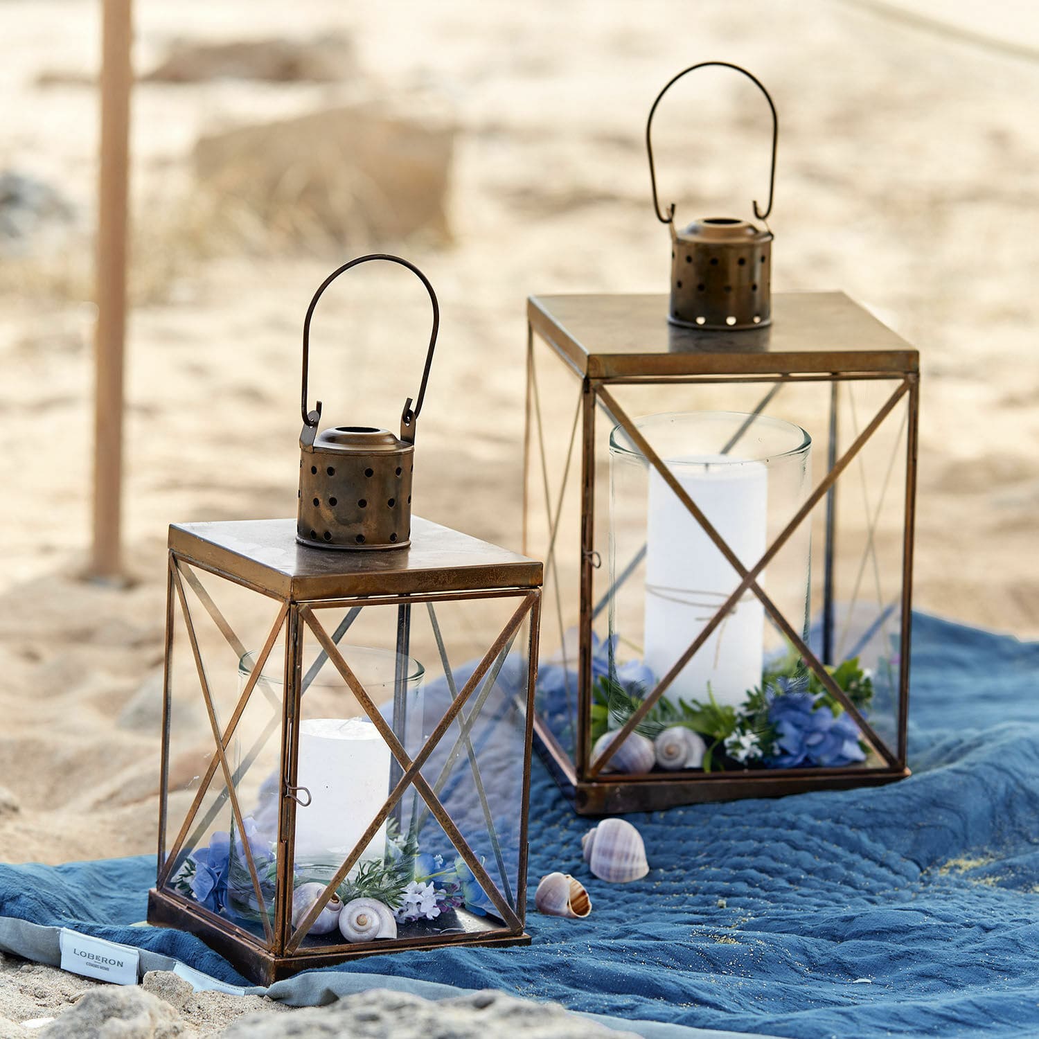 Laterne 2er Set Garance Zwei braune Metalllaternen mit weißen Kerzen und Dekorationen auf blauem Tuch am Strand