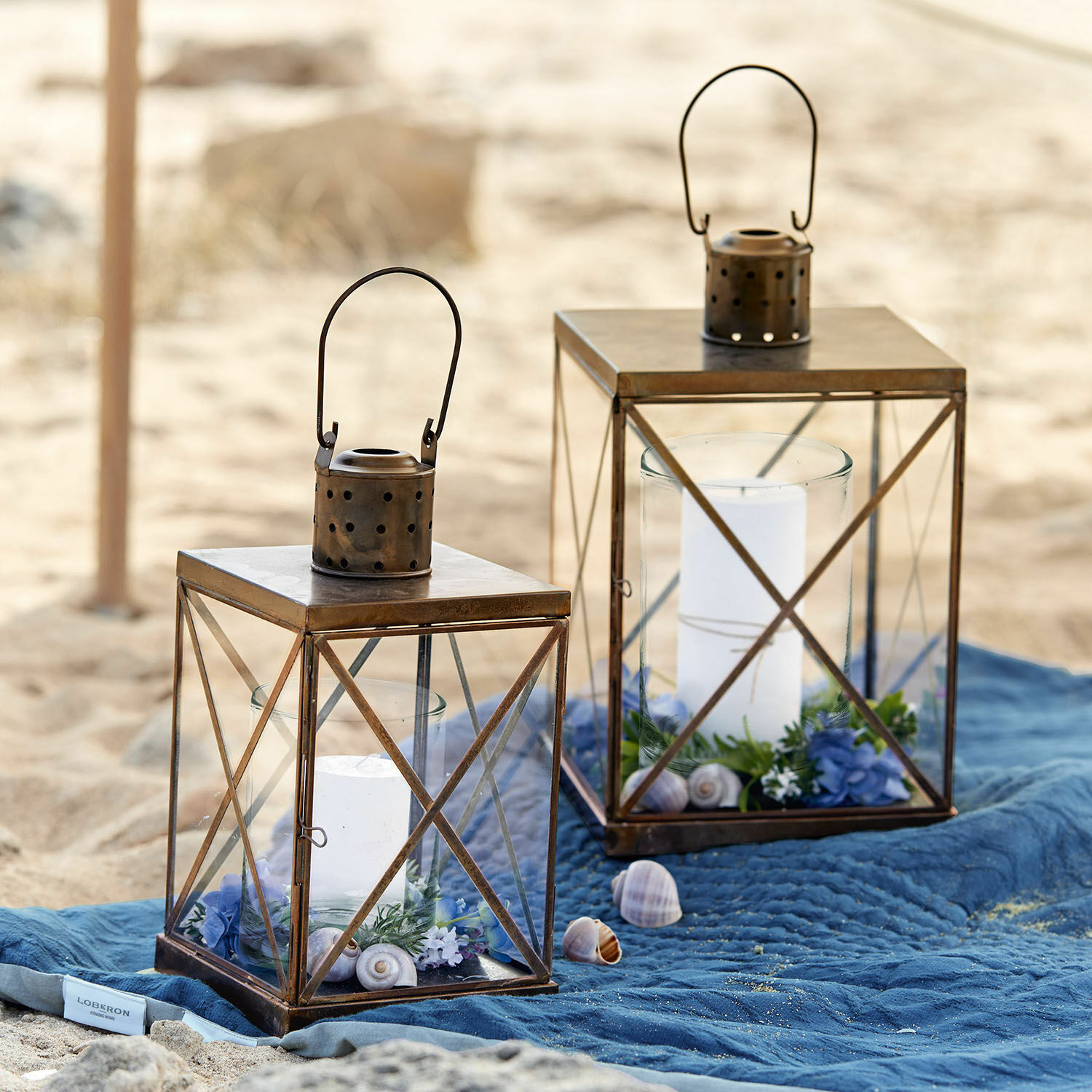 Laterne 2er Set Garance Zwei braune Metalllaternen mit weißen Kerzen und Dekorationen auf blauem Tuch am Strand