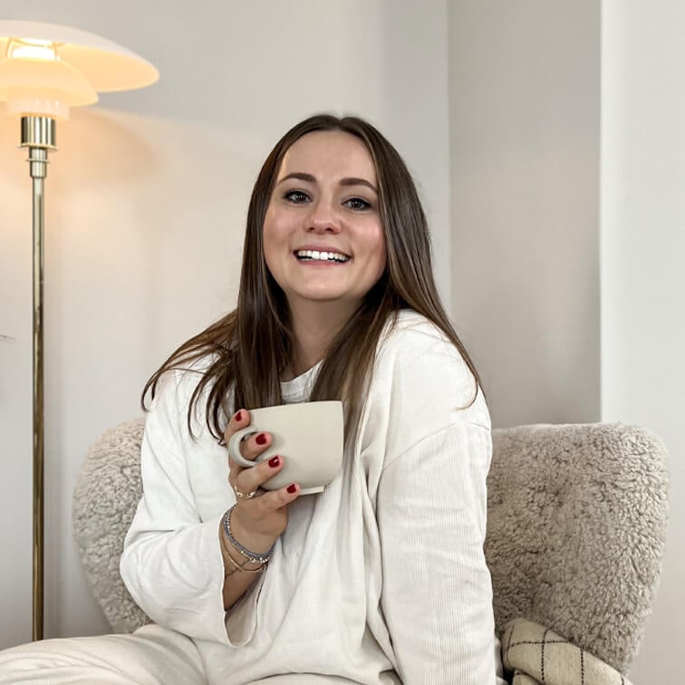 Lächelnde Frau im weißen Pullover hält eine beige Tasse und sitzt in einem gemütlichen Sessel im warmen Raum
