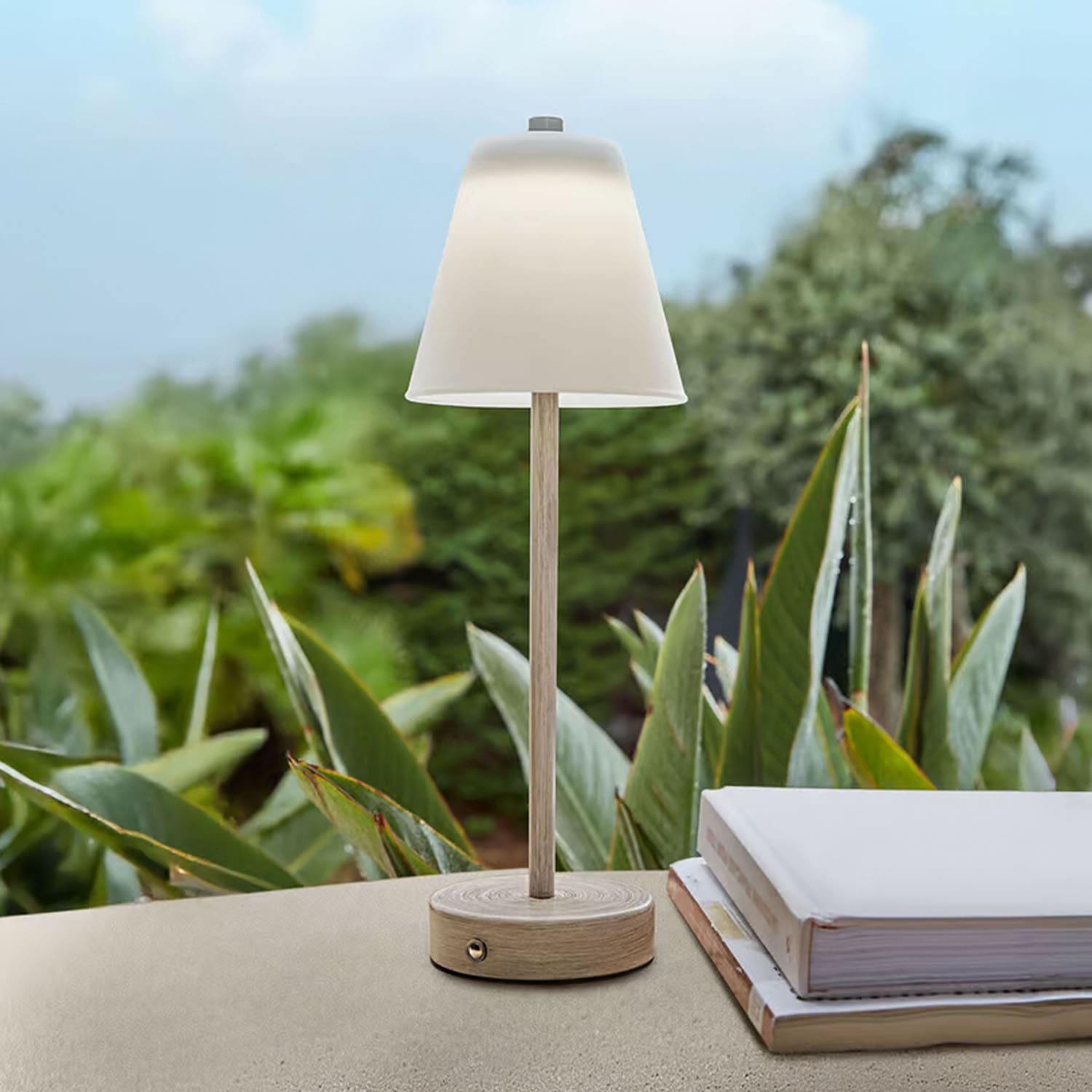 Tischlampe aus hellem Holz mit weißem Lampenschirm vor grünen Pflanzen, Wohnaccessoire