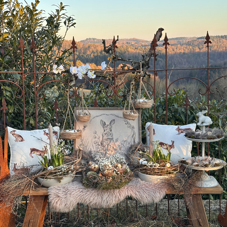 Rustikale Osterdekoration mit Hasen-Kissen, Weidenkörben und natürlichen Blumen auf einer Holzbank