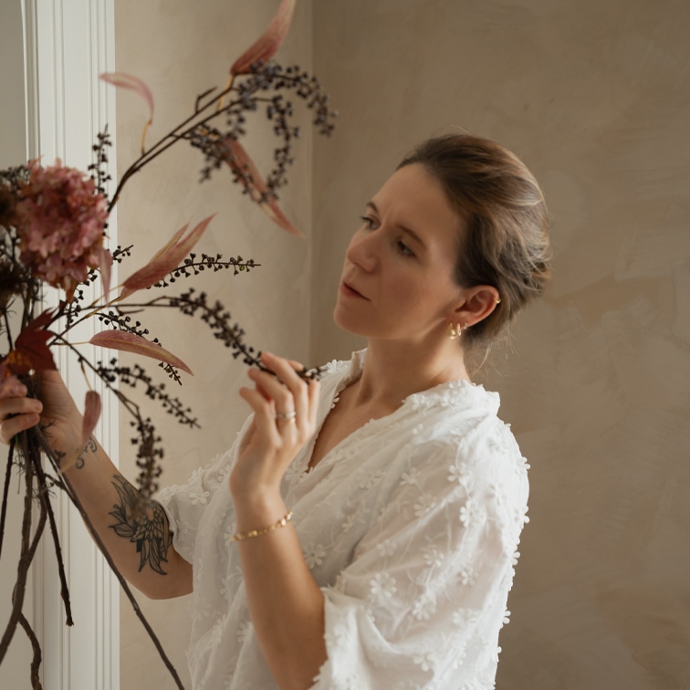 Frau in weißer Spitzenbluse arrangiert getrocknete Blumendekoration in hellem Raum