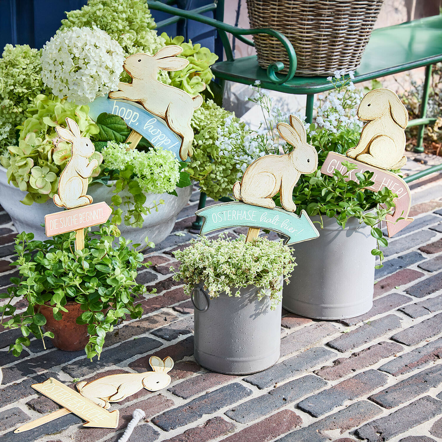 Holz-Osterdekorationen mit Hasen und Schriftzügen in Pflanzgefäßen mit grünen Pflanzen auf gepflastertem Boden