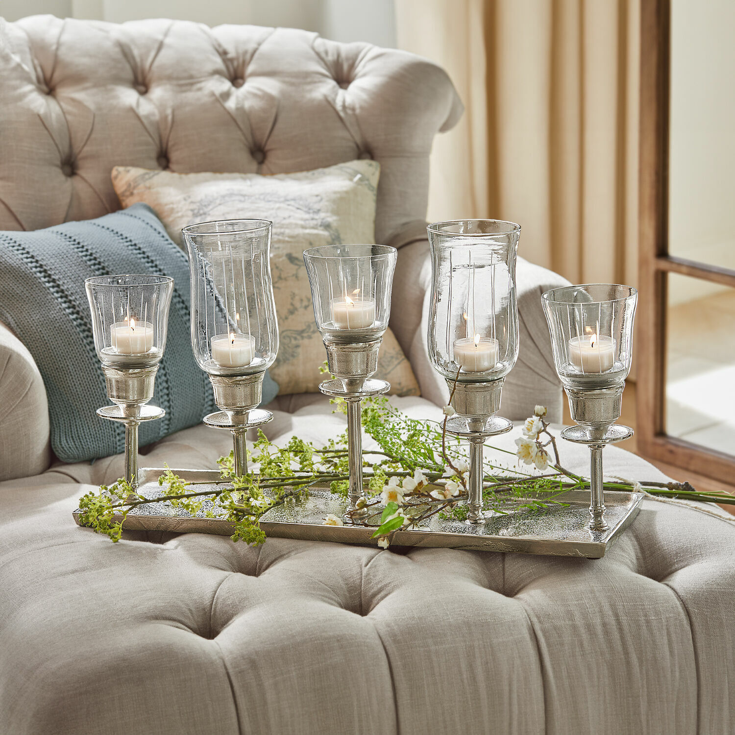 Kerzentablett Ennedi Fünf silberne Kerzenhalter aus Glas und Metall auf beigem Sofa mit Kissen arrangiert