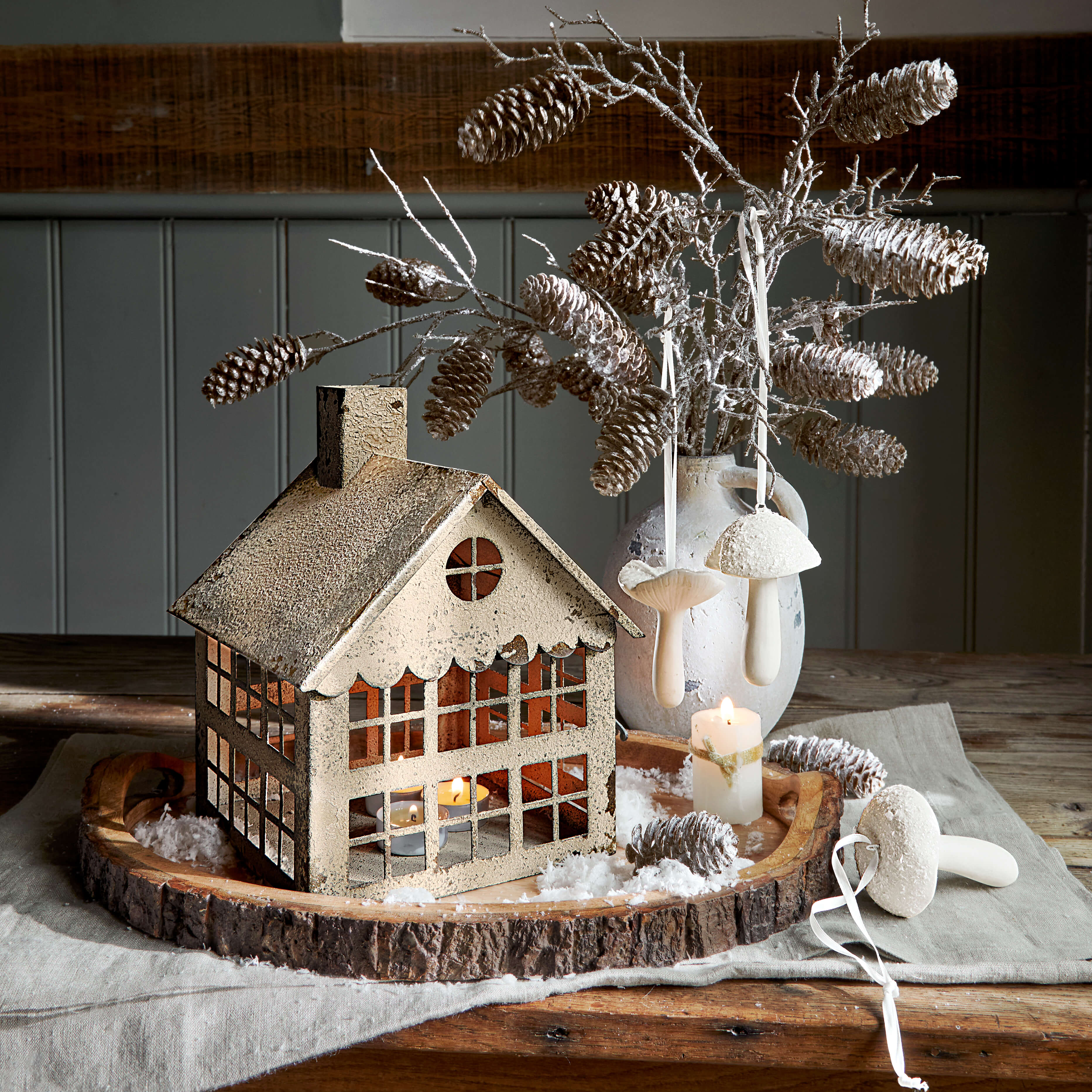 Dekoratives Metallhaus mit Teelicht auf Holzplatte, Tannenzapfen und weißen Pilzanhängern