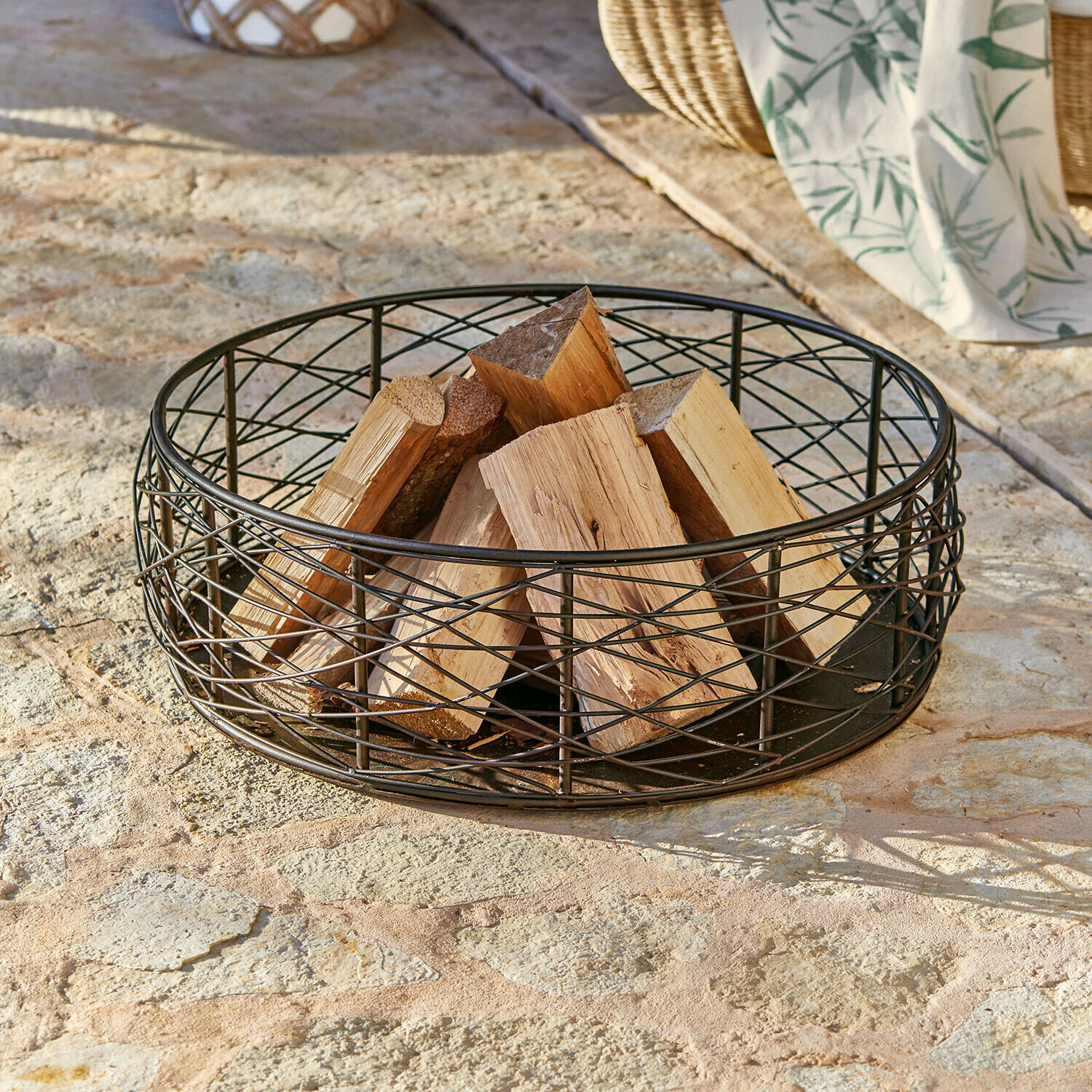 Schwarzer Metallkorb mit naturbelassenem Brennholz auf Steinboden für Kaminholzlagerung