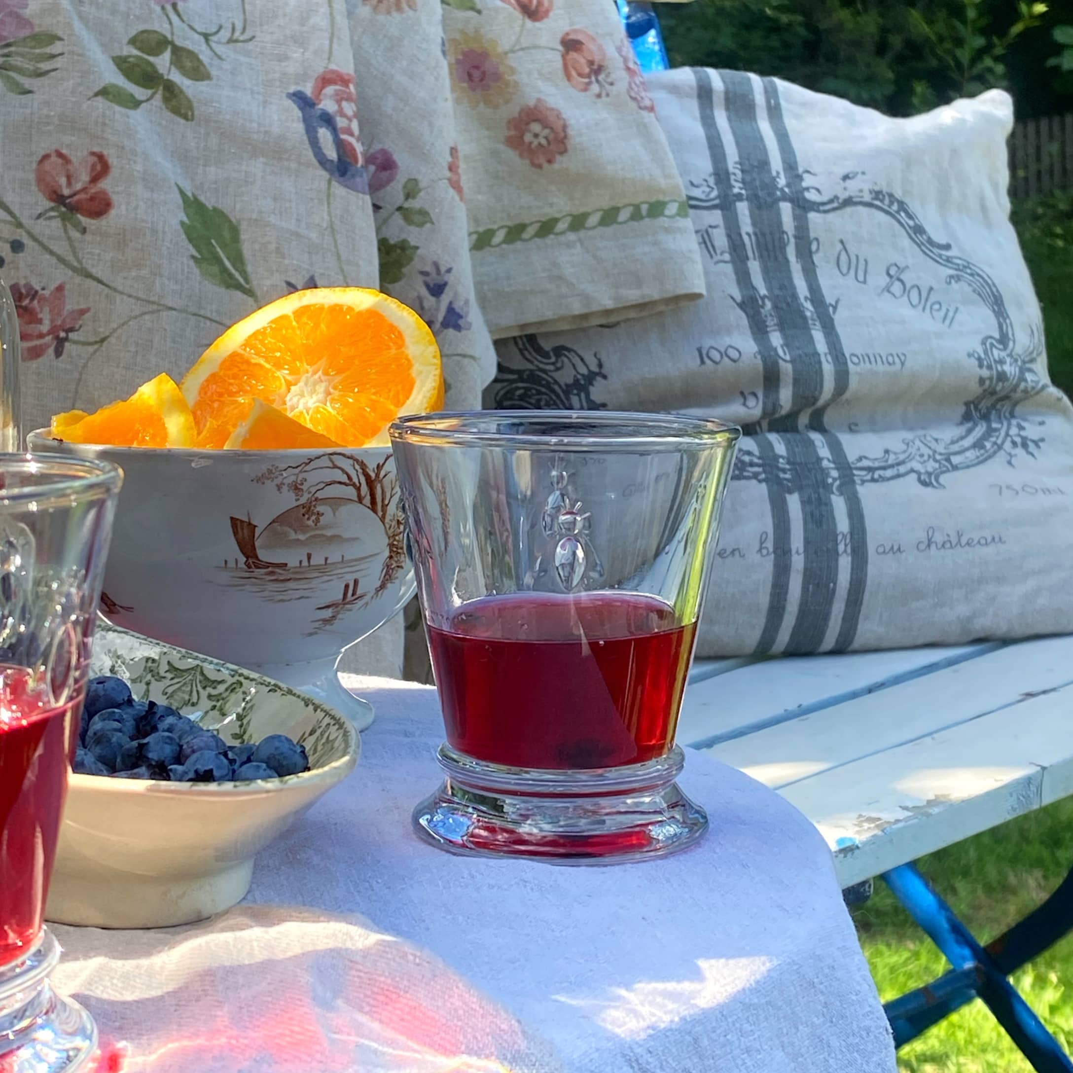 Glas mit rotem Getränk auf weißem Tuch mit Früchten und Kissen im Garten