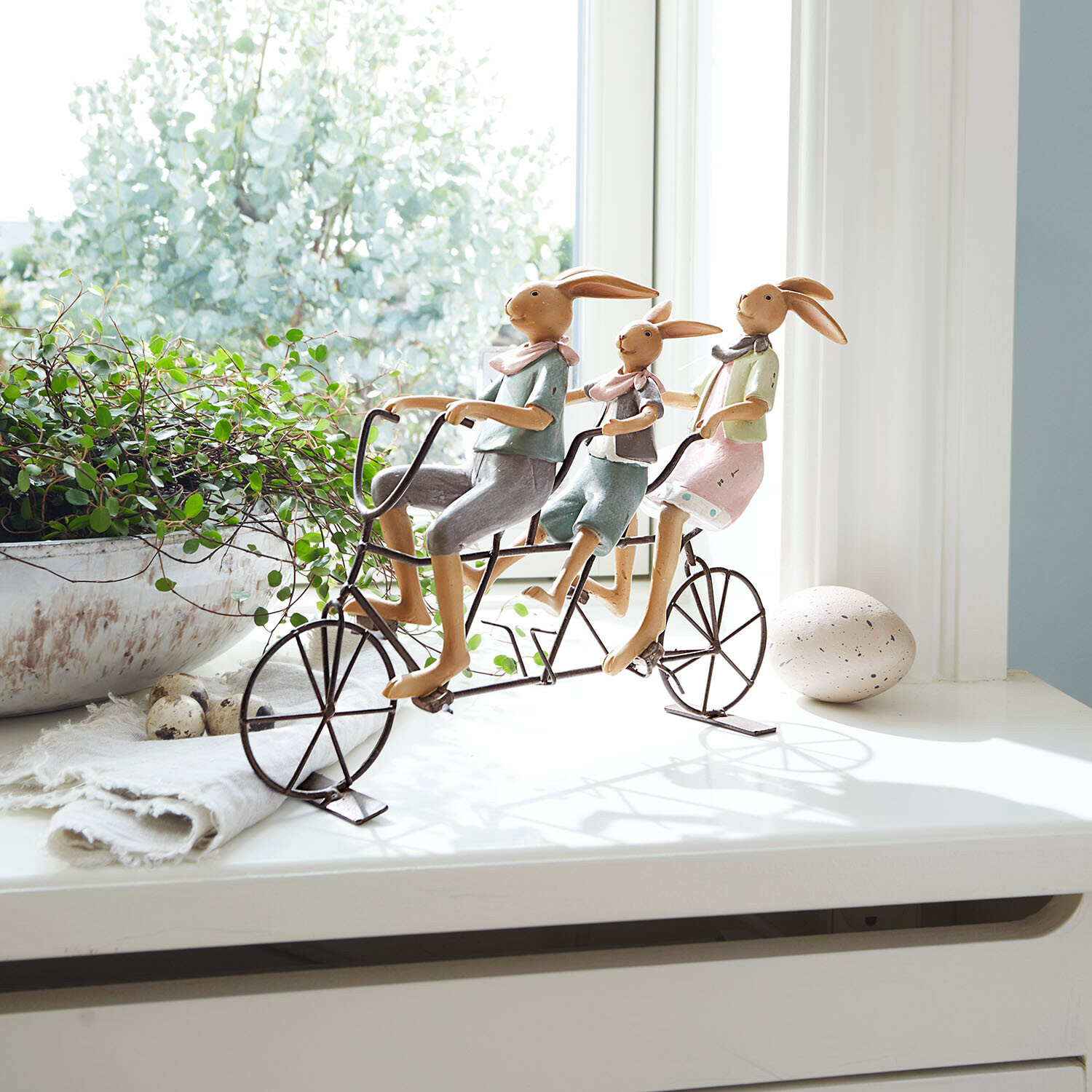 Dekorative Hasenfamilie aus Metall auf pastellfarbenem Fahrrad auf Fensterbank mit Pflanze
