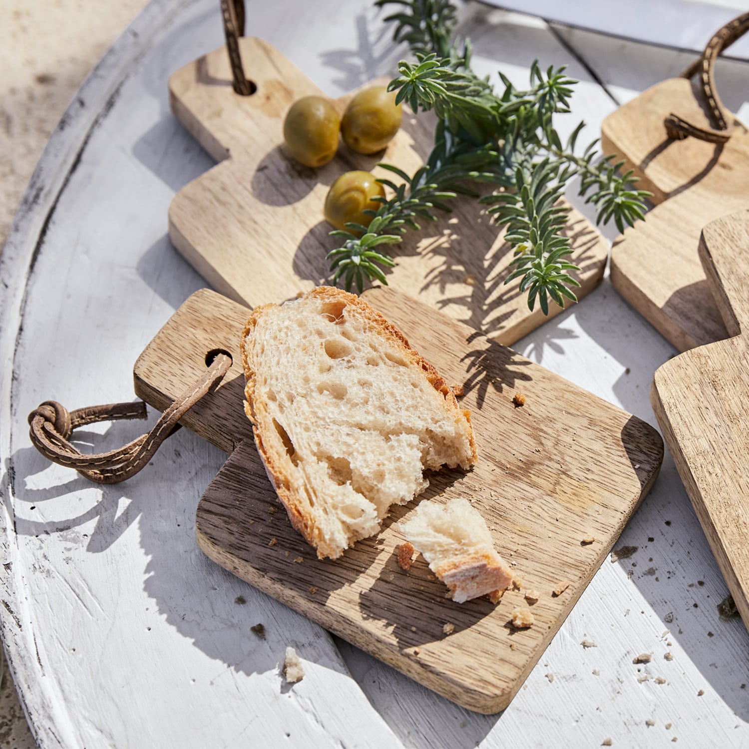 Holz-Schneidebrett mit Brotscheibe, grünen Oliven und Kräutern auf weißem Tisch