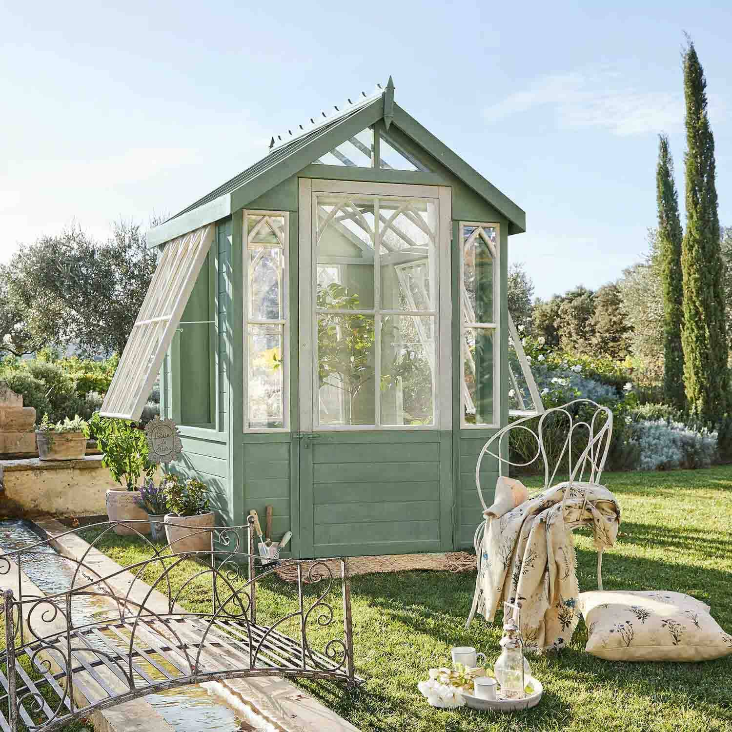 Grünes Holz-Gewächshaus mit weißen Fensterrahmen im Garten, Metallstuhl mit Kissen daneben