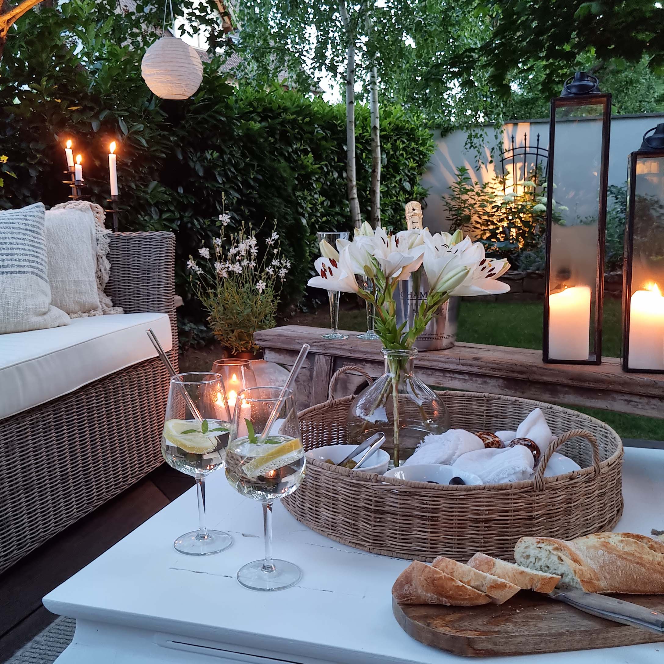 Gemütlicher Garten mit Korbsofa, weißen Lilien und Laternen auf weißem Holztisch