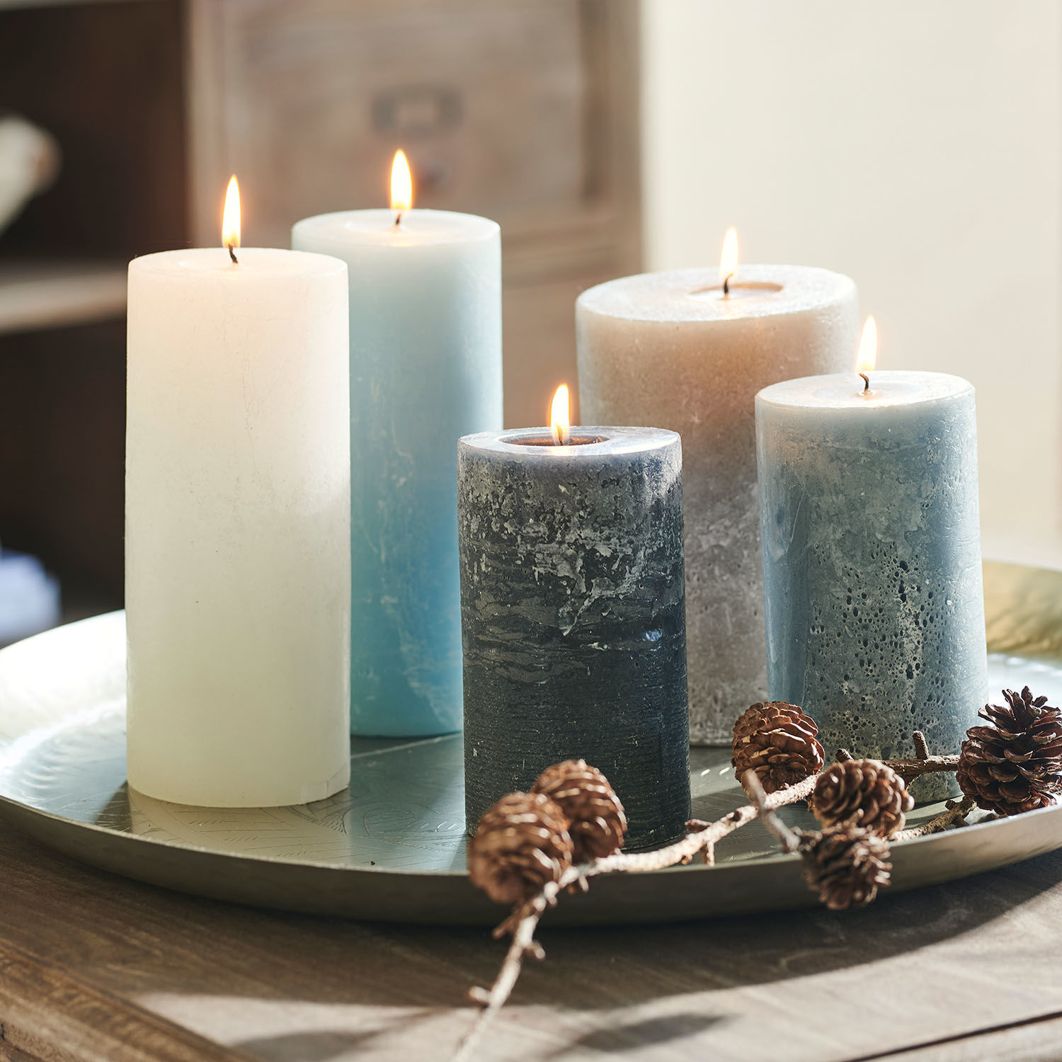 Fünf Kerzen in verschiedenen Blau- und Weißtönen auf Metalltablett mit Tannenzapfen dekoriert